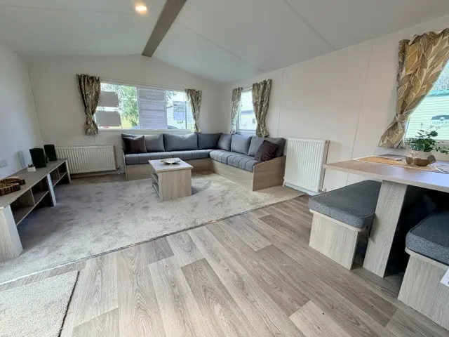 Modern living area with grey sectional sofa, coffee table, dining nook, and large windows with curtains. Light wood flooring and neutral décor create a bright, open space.